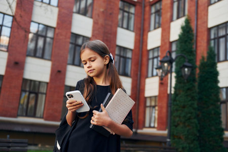 Holding phone in hands. Schoolgirl is outside near school buildingの写真素材
