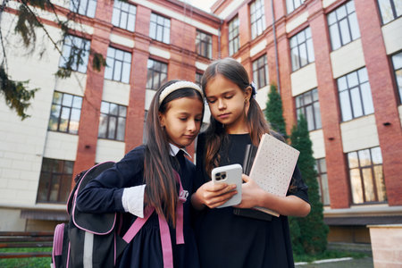 With books. Two schoolgirls is outside together near school buildingの写真素材