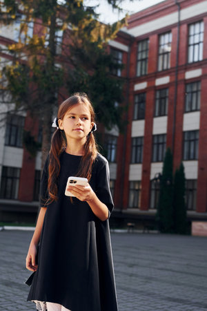 In black uniform. Schoolgirl is outside near school buildingの写真素材