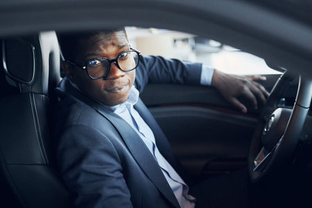 In glasses. Young african american businessman in black suit is in the automobile.の写真素材