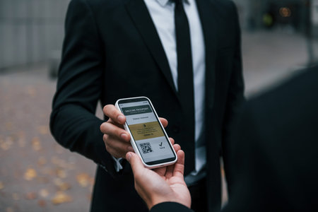 Holding phone with vaccination certificate. Businessman in black suit and tie is outdoors in the...