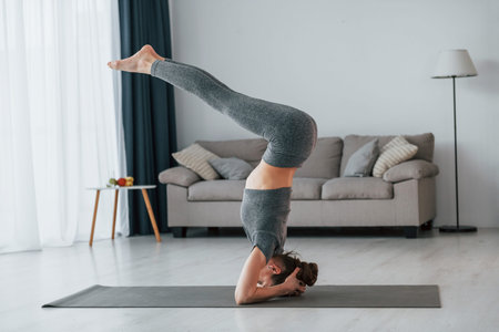 Doing headstand. Young woman with slim body type and in yoga clothes is at home.