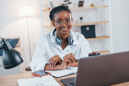 With laptop on the table. Young african american woman in white shirt is at home.