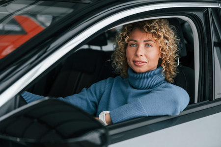 Sitting at driver's seat. Woman with curly blonde hair is in autosalon.
