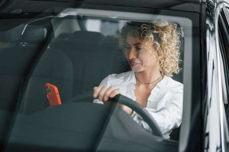 Using phone. Woman with curly blonde hair is in autosalon.