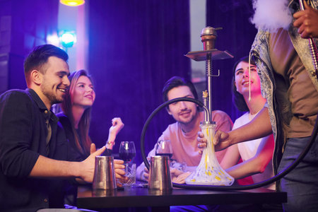 Talking with each other when sitting by the table with hookah. Group of friends having fun in the night club together.の写真素材