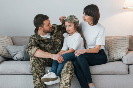 On the sofa. Soldier in uniform is at home with his wife and daughter.
