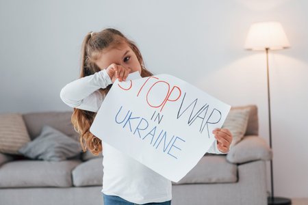 Little girl standing at home and holding banner with stop war in Ukraine text.