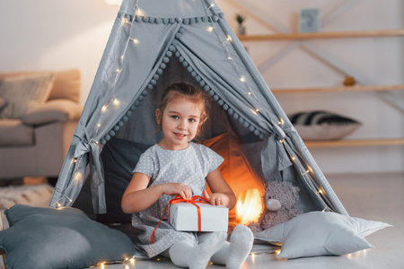 Holding gift box. Cute little girl playing in the tent that is in the domestic room.