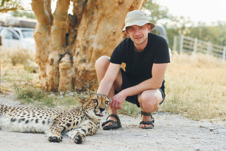 Male tourist in casual clothes and hat sitting and petting a cheetah outdoors.の写真素材