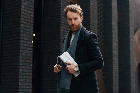 Holding newspaper in the hand. Stylish man with beard is outdoors near building.の写真素材