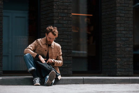 Sitting on the sidewalk. Stylish man with beard in khaki colored jacket and in jeans is outdoors...