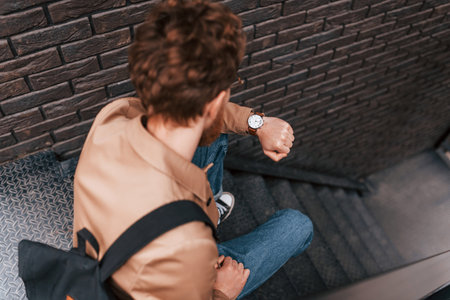 Sitting and checking time. Man in khaki colored jacket and in jeans is on the stairs on the building...