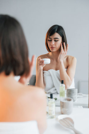 Sitting by the mirror and using cream on face. Young woman is at home. Conception of beauty.の写真素材