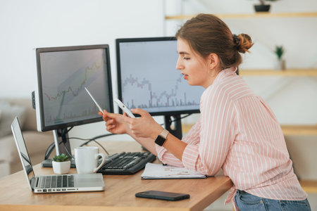 Using smartphone. Female stock broker in casual clothes is working in the office by pc.