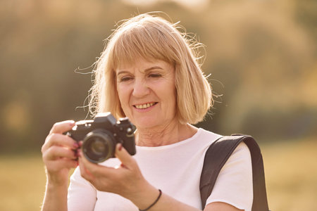 Using the camera. Making the photos. Senior woman having nice weekend outdoors on the field at sunny...