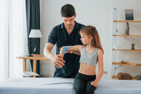 Girl is sitting. Professional physiotherapist putting kinesio tape on kid arms in clinic.