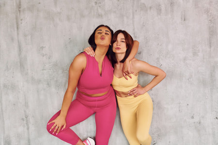 Standing and embracing each other. Beautiful women in sportive clothes is indoors in the studio.