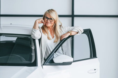 Leaning on the automobile. Woman in white formal clothes is in the car dealership.
