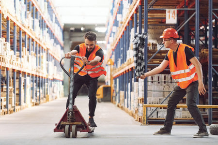 Man riding trolley platform. Having fun. Two storage workers is in the warehouse.