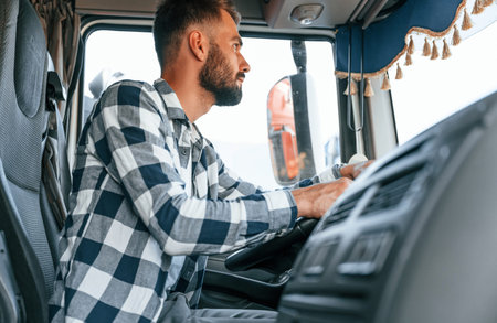 Sitting in the cabin by steering wheel. Young truck driver is with his vehicle at daytime.