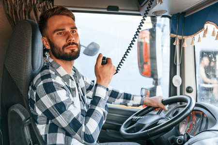 Using radio. Young truck driver is with his vehicle at daytime.