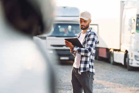 With black notepad. Young truck driver is with his vehicle at daytime.