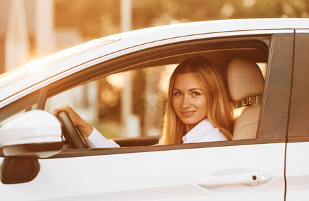 Sitting and driving. Young woman in white clothes is with her electric car at daytime.