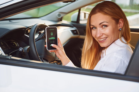 Sitting inside with smartphone. Young woman in white clothes is with her electric car at daytime.