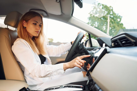 Navigating the vehicle's interface. Young woman in white clothes is with her electric car at...