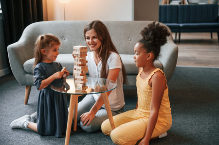 Sitting on the floor. A young woman with two girls is playing a wooden tower game indoors together.