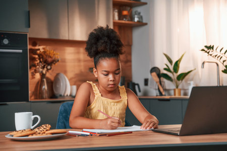 Sitting by the table with laptop and paper for drawing. Cute black girl in casual clothes is at home...