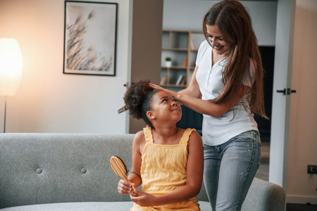 Mother is combs hair. Young woman with her daughter is together at apartment.