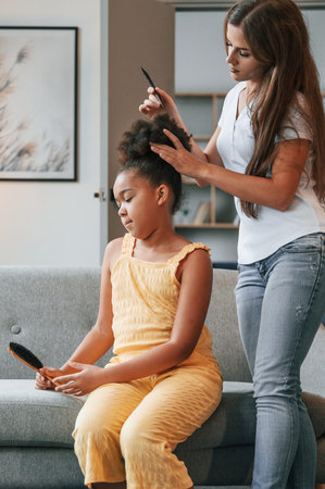 Mother is combs hair. Young woman with her daughter is together at apartment.の写真素材