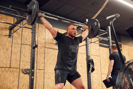 Trying hard facial expression. Strong man is lifting barbell in the gym.