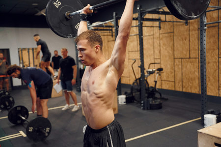 Standing with hands straight up. Strong man is lifting barbell in the gym.