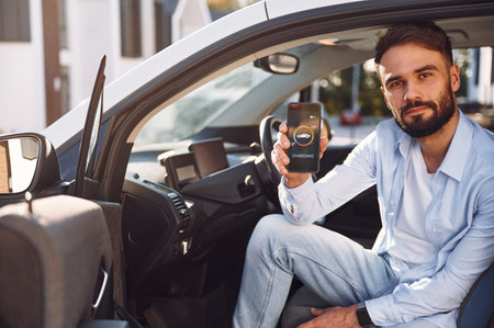 Sitting inside the vehicle, monitoring charging process by using smartphone. Young stylish man is...