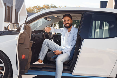 Sitting in the vehicle and smiling. Young stylish man is with electric car at daytime.
