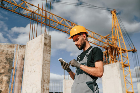 With phone in hands. Standing against crane. Man in uniform is working on the construction site.