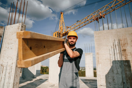 Holding and walking with wooden plank. Man in uniform is working on the construction site.