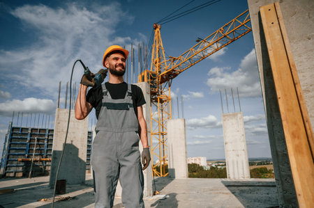 Standing and holding drill. Man in uniform is working on the construction site.