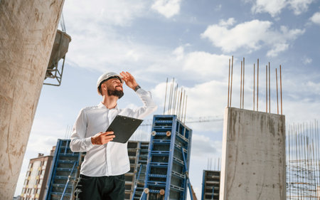 Engineer is holding notepad. Man in uniform is working on the construction site.