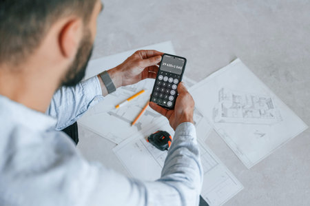 Calculating numbers by using smartphone. Plan is on the floor. The man is making repairs in the...