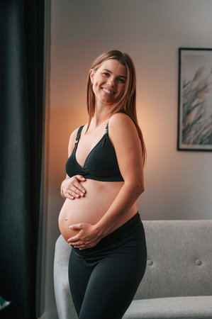 Standing and having positive mood. Beautiful pregnant woman is indoors at home.