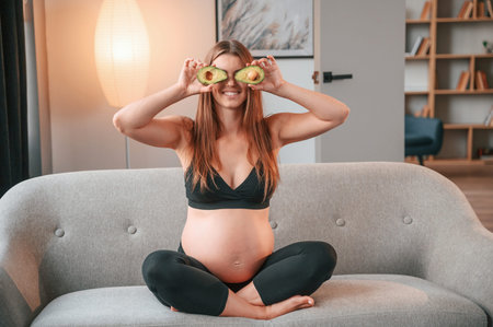 Holding avocado. Beautiful pregnant woman is indoors at home.
