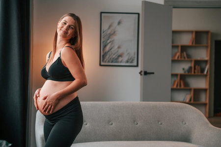 Standing and having positive mood. Beautiful pregnant woman is indoors at home.