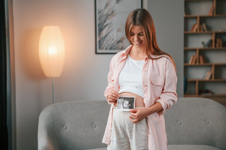 Holding ultrasound diagnostics picture of the baby. Beautiful pregnant woman is indoors at home.