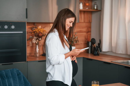 Using smartphone. Beautiful pregnant woman is indoors at home.