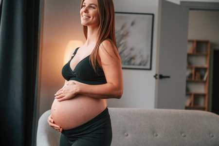 Standing near the sofa and smiling. Beautiful pregnant woman is indoors at home.