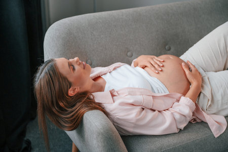 Resting on the sofa. Beautiful pregnant woman is indoors at home.の写真素材